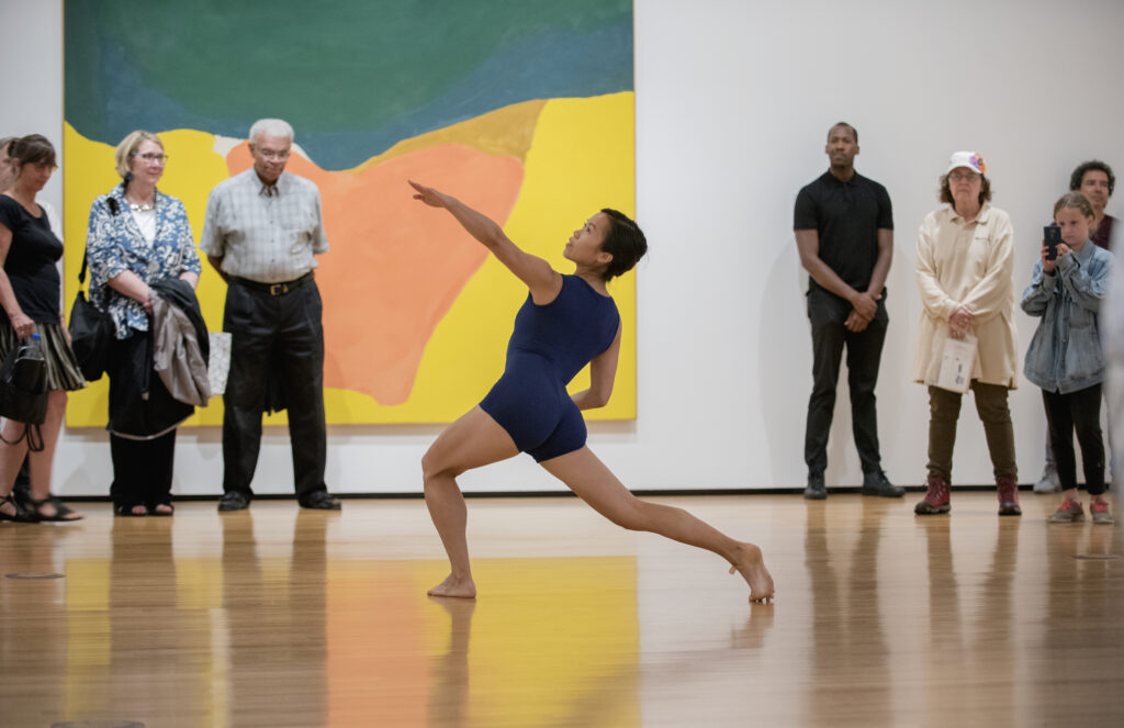 A woman in a dark blue leotard dances in a museum gallery.